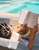 a person lying on a lounge chair reading a book by a pool